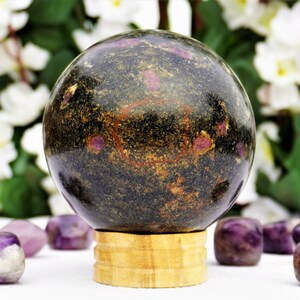 Può includere: Una sfera nera e dorata con macchie rosa, appoggiata su un supporto in legno. La sfera è circondata da pietre di ametista viola.