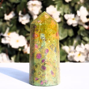 May include: A green and pink crystal point with a pointed top and a flat base. The crystal has a speckled appearance with green, pink, and gold flecks.
