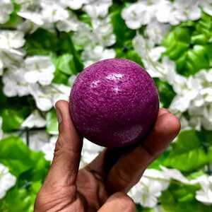 May include: A deep purple sphere with a textured surface. The spherical object is held in a hand against a backdrop of white flowers and green foliage. The sphere's colour is a rich, saturated hue.