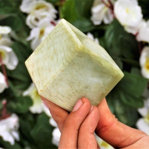 Può includere: Un cristallo di forma cubica di colore verde pallido con una superficie liscia e lucida. Il cristallo è tenuto nelle mani di una persona.