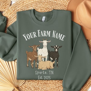 May include: Green sweatshirt featuring a graphic of four goats and the text "YOUR FARM NAME", "Sparta, TN", and "Est. 2025". The goats are brown, white, and black. The sweatshirt is folded on a woven surface.