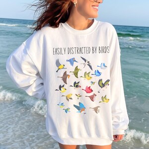May include: White sweatshirt with the text "Easily Distracted by Birds" and a graphic of colorful birds flying around.