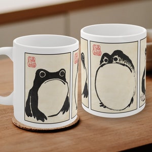May include: White ceramic coffee mug featuring a black and white frog illustration in a Japanese art style. The frog is depicted in a series of poses within rectangular frames. The mug sits on a wooden coaster.