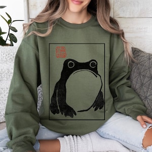 May include: Olive green sweatshirt featuring a black frog illustration within a black rectangular frame. The frog has large eyes and a prominent mouth. Above the frog is a red Japanese seal. The sweatshirt is a casual garment.