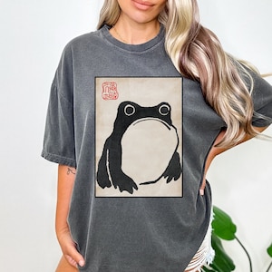Op de afbeelding: Een donkergrijs T-shirt met een vintage-stijl illustratie van een kikker in een rood kader. De kikker is zwart-wit en heeft een grote, ronde buik. De tekst "蛙" staat in rood in de linkerbovenhoek van het kader.