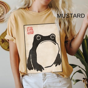 Op de afbeelding: Een mosterdgeel T-shirt met een zwart-witte illustratie van een kikker. De kikker bevindt zich in een vierkant frame met een rode stempel waarop "蛙" in het Japans staat.