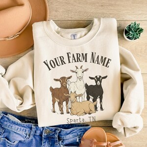 May include: Cream-colored sweatshirt with a graphic of four goats and the text "YOUR FARM NAME" above them. Below the goats is the text "Sparta, TN". The sweatshirt is paired with denim shorts, a hat, and sandals.