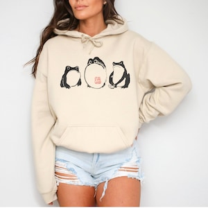 May include: A cream-colored hoodie with a black and white graphic of three frogs. The frogs are in a line, with the middle frog being the largest. The middle frog has a red stamp on its belly that says "The Great Frog".