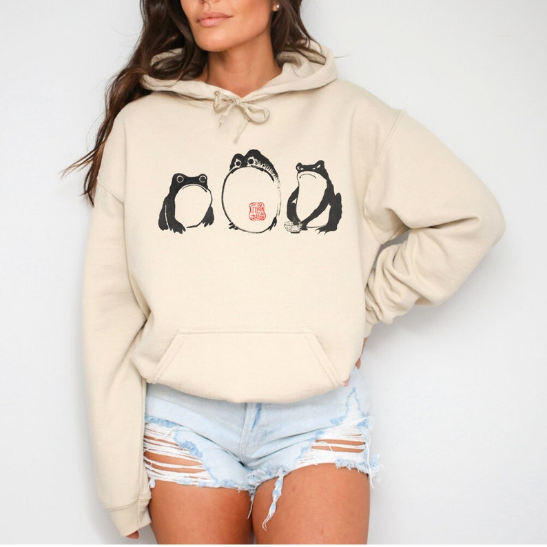 May include: A cream-colored hoodie with a black and white graphic of three frogs. The frogs are in a line, with the middle frog being the largest. The middle frog has a red stamp on its belly that says "The Great Frog".