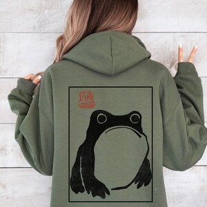 May include: A green hooded sweatshirt with a black and white frog illustration on the back. The frog is in a square frame with a red stamp in the top left corner.