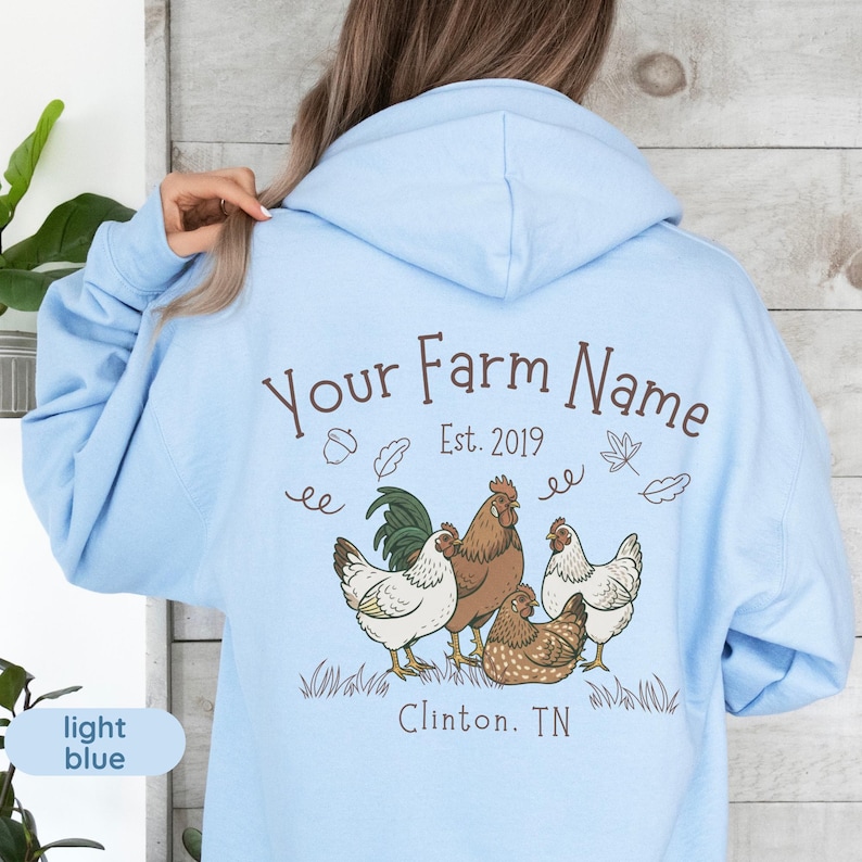 May include: Light blue hoodie with a farm-themed graphic. The design features a rooster and several chickens, along with the text "Your Farm Name Est. 2019" and the location "Clinton, TN". The hoodie is a soft, casual garment.