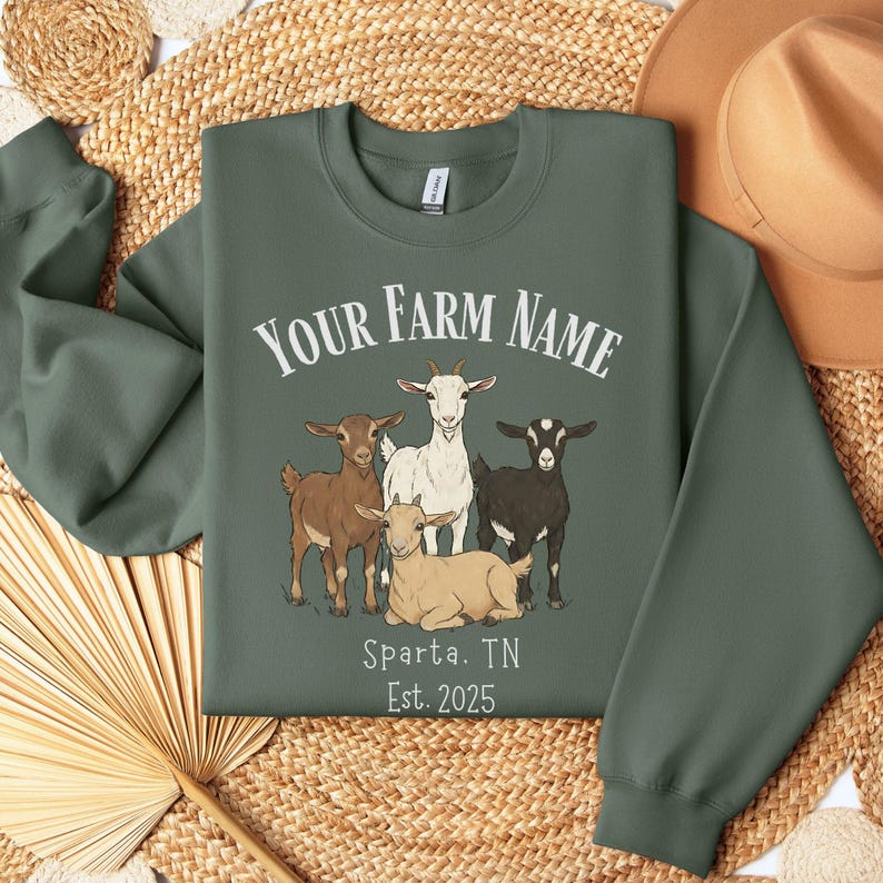 May include: Green sweatshirt featuring a graphic of four goats and the text "YOUR FARM NAME", "Sparta, TN", and "Est. 2025". The goats are brown, white, and black. The sweatshirt is folded on a woven surface.
