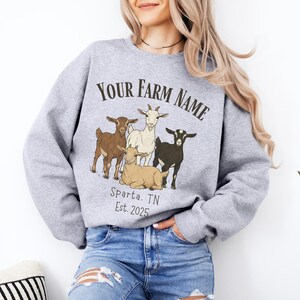 May include: A light gray sweatshirt featuring a graphic of four goats in brown, white, and black. The text "YOUR FARM NAME" is arched above the goats, with "Sparta, TN Est. 2025" below. The sweatshirt is a crew neck style.