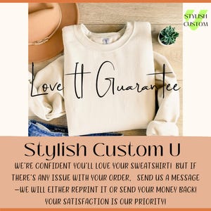 May include: Cream-colored sweatshirt with the text "Love It Guarantee" in black script. The sweatshirt is folded on top of blue jeans. A brown hat and a small succulent plant are also visible. The text "Stylish Custom U" is at the bottom.