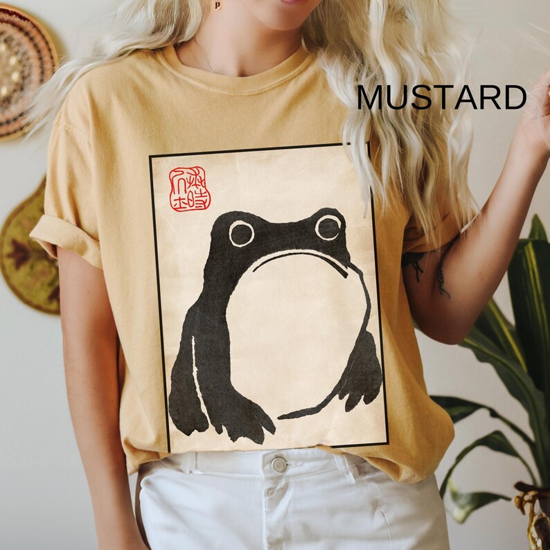 Op de afbeelding: Een mosterdgeel T-shirt met een zwart-witte illustratie van een kikker. De kikker bevindt zich in een vierkant frame met een rode stempel waarop "蛙" in het Japans staat.