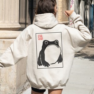 May include: A cream-colored hoodie with a black and white illustration of a frog on the back. The frog is in a square frame with a red stamp in the top left corner.