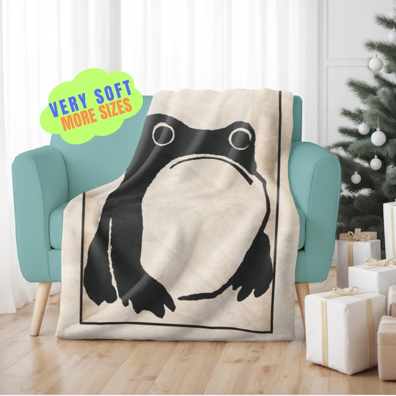 May include: A soft, beige throw blanket with a black and white illustration of a frog. The frog has large, round eyes and a simple, cartoon-like design.