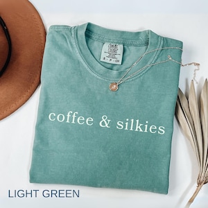 May include: Light green t-shirt with the text "coffee & silkies" in white. A gold necklace with a pendant rests on the shirt. The shirt is folded on a white surface, with a brown hat and dried plant stems in the background.