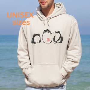 May include: A cream-colored hoodie with a black and white illustration of three frogs. The frogs are sitting in a row and the middle frog has a red stamp on its back. The hoodie has a drawstring hood and a kangaroo pocket.