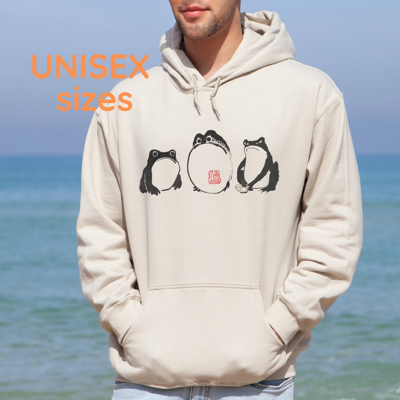 May include: A cream-colored hoodie with a black and white illustration of three frogs. The frogs are sitting in a row and the middle frog has a red stamp on its back. The hoodie has a drawstring hood and a kangaroo pocket.