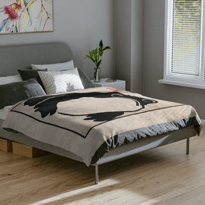 May include: A beige throw blanket with a black and white graphic design of a stylized animal. The blanket is on a bed with gray bedding and pillows.