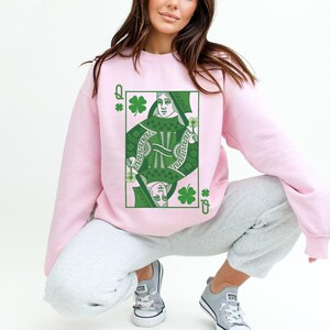May include: A pink sweatshirt with a green Queen of Clubs playing card graphic. The card features a woman holding a shamrock.