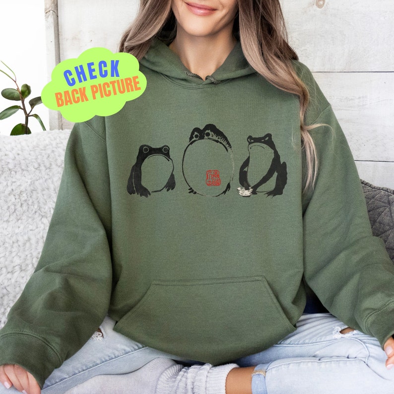 May include: A green hooded sweatshirt with a graphic of three frogs and a red stamp. The frogs are in black and white and the stamp is in red. The sweatshirt is a casual style and is perfect for everyday wear.