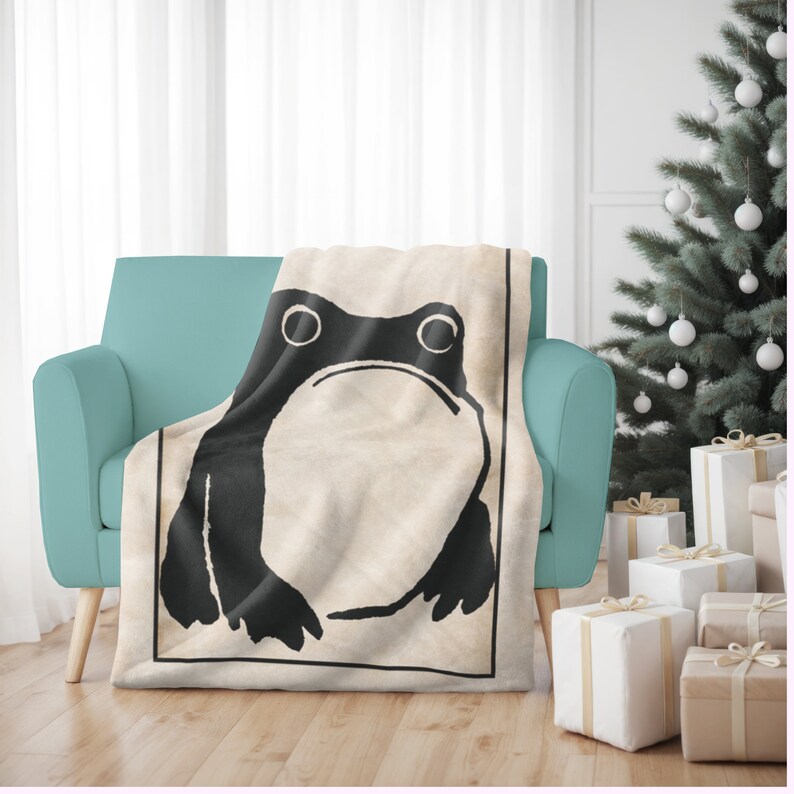 May include: A cream-colored throw blanket with a black and white illustration of a frog. The blanket is on a teal blue armchair.