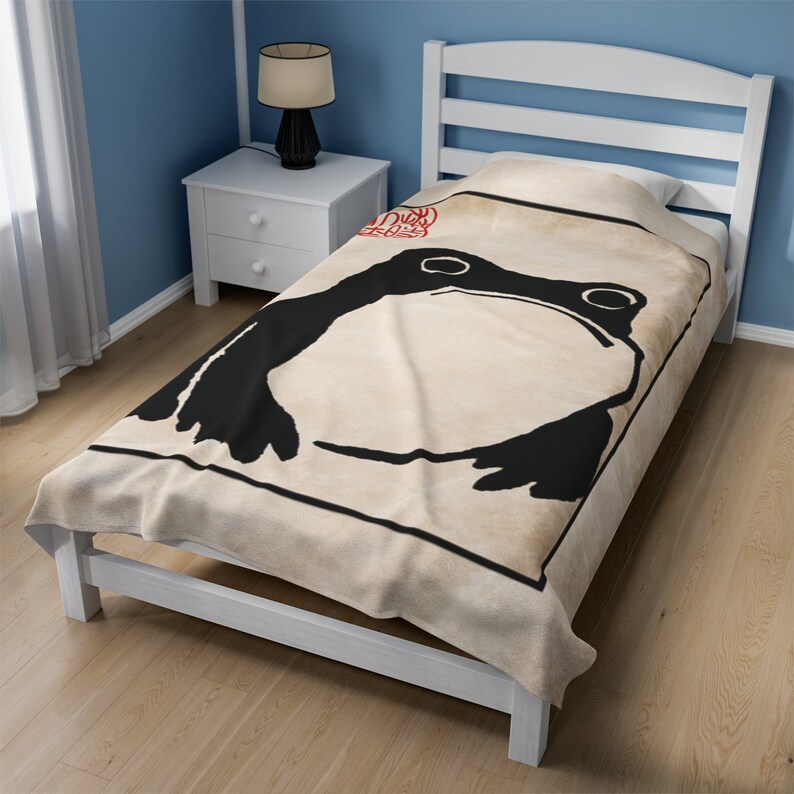 May include: A beige fleece throw blanket with a black and white illustration of a frog. The frog is in a square frame with a red stamp on the top right corner.