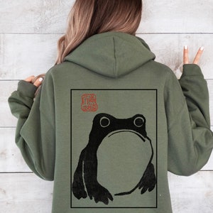 May include: A green hooded sweatshirt with a black and white frog illustration on the back. The frog is in a square frame with a red stamp in the top left corner.