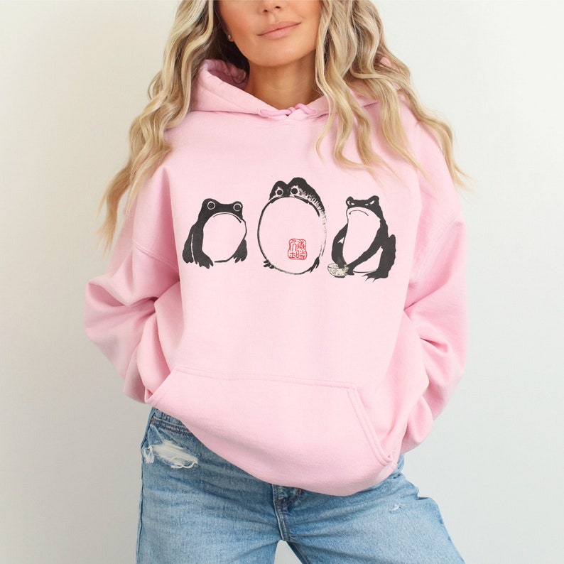 May include: A pink hoodie with a graphic of three frogs. The frogs are black and white with a red stamp on the middle frog. The stamp says "Good Luck".