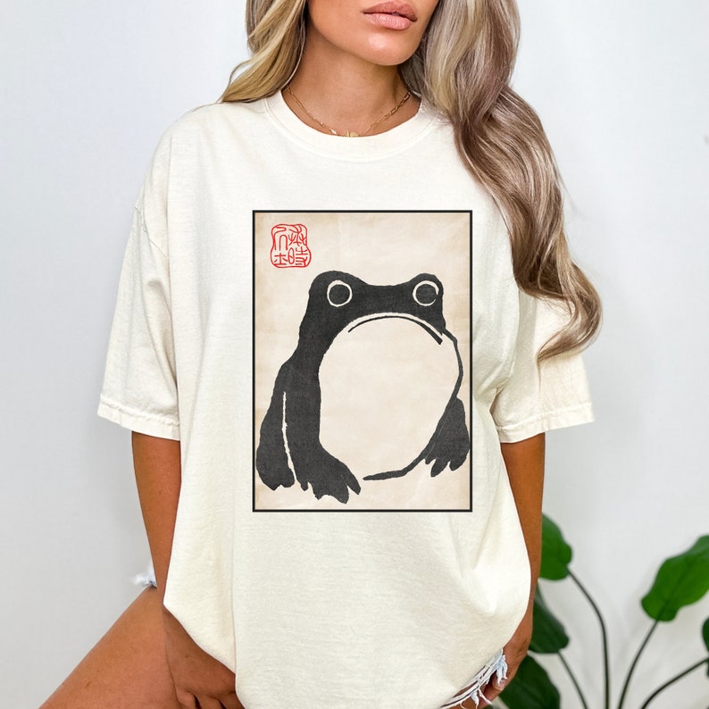 Op de afbeelding: Een wit T-shirt met een zwart-witte illustratie van een kikker in een rood vierkant met Japanse karakters. De kikker kijkt recht naar de kijker.