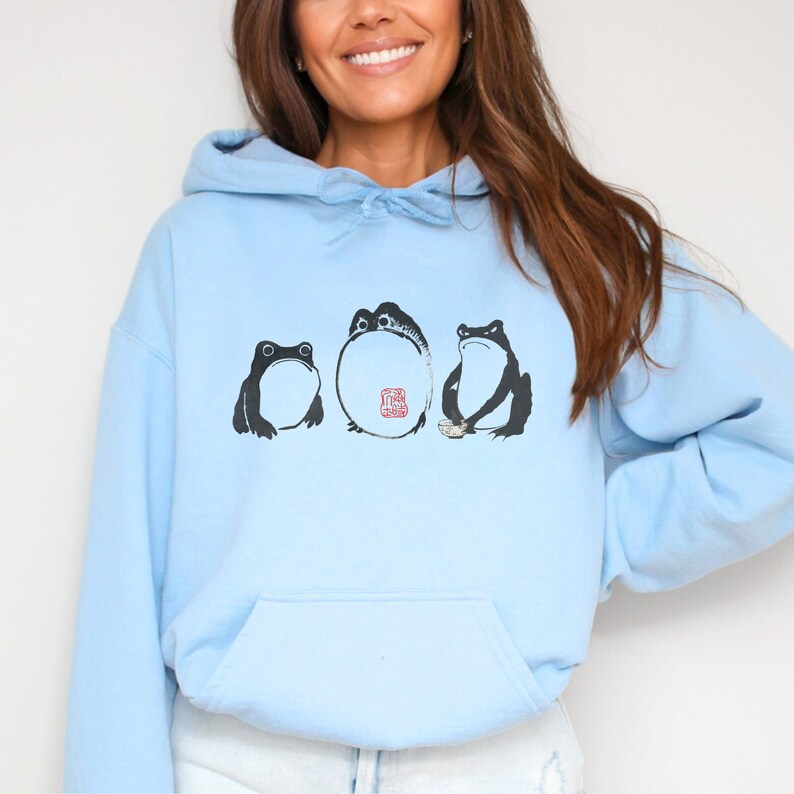 May include: A light blue hoodie with a graphic of three frogs. The frogs are black and white and are arranged in a row. The frog in the middle is larger than the other two and has a red seal on its belly.