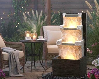 Könnte beinhalten: Eine Garten-Terrasse bei Dämmerung mit einem mehrstufigen Wasserspiel, Korbstühlen und einem kleinen Tisch. Lichterketten und Kerzen sorgen für Beleuchtung, mit Topfblumen, die Farbe hinzufügen. Eine gemütliche, einladende Atmosphäre.
