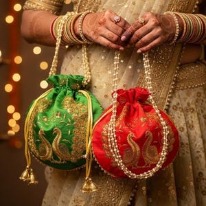 May include: Two drawstring pouches, one green and one red, featuring gold embroidered peacock motifs. Each bag has a pearl-beaded handle and gold drawstring cords with metallic tassels. The fabric has a satin-like finish with intricate gold thread detailing.