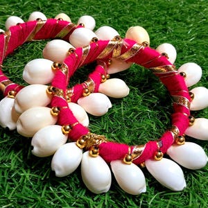 Puede incluir: Dos brazaletes de tela rosa y dorada decorados con conchas blancas. Los brazaletes están sobre una superficie de hierba verde.