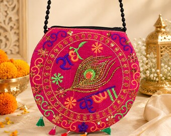 Puede incluir: Un bolso bandolera redondo de color rosa intenso con un intrincado bordado de una pluma de pavo real y escritura devanagari azul. El bolso tiene una correa de cuentas negras, cierre de cremallera y borlas de tela coloridas en el borde.