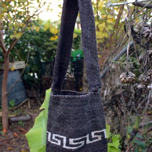 Puede incluir: Una bolsa de mano de punto gris con un dise&ntilde;o geom&eacute;trico blanco. La bolsa tiene una correa larga y est&aacute; colgada de una rama.