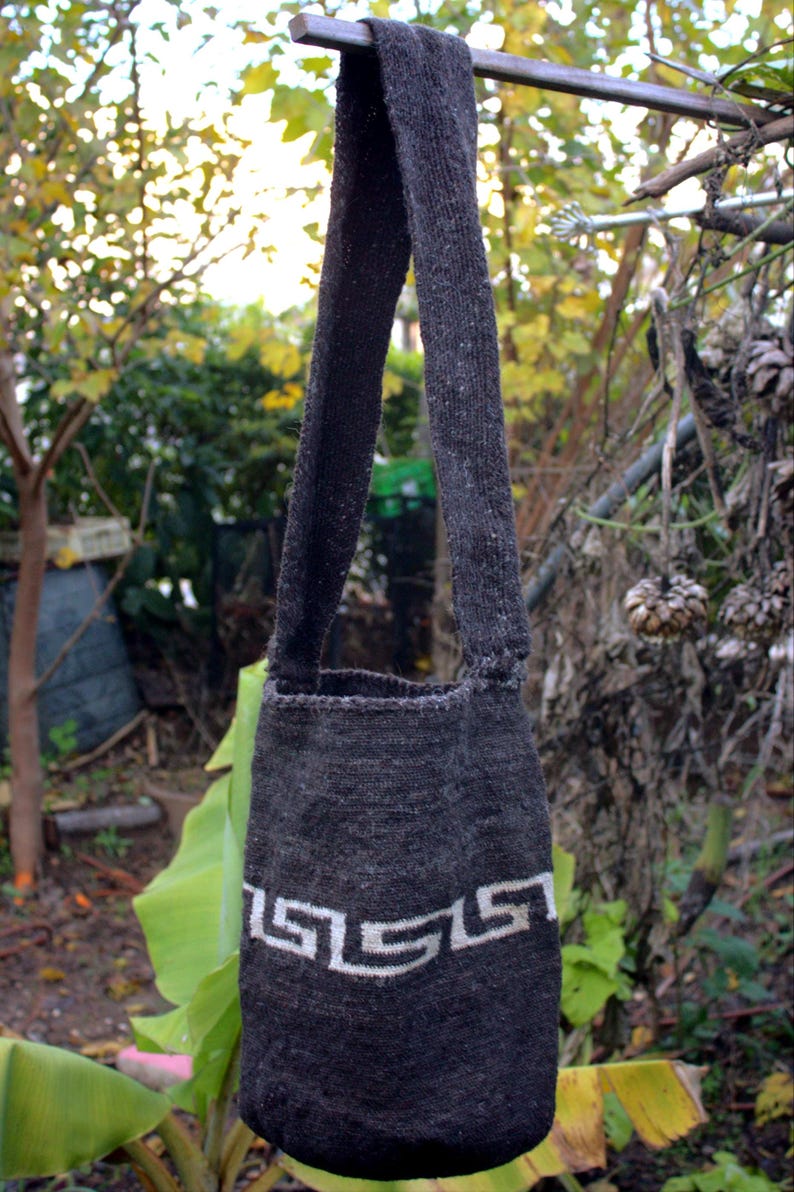 Puede incluir: Una bolsa de mano de punto gris con un dise&ntilde;o geom&eacute;trico blanco. La bolsa tiene una correa larga y est&aacute; colgada de una rama.