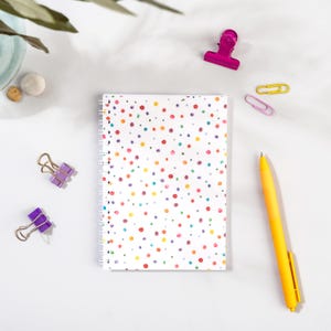 May include: A white spiral-bound notebook featuring a pattern of colourful polka dots. A yellow pen and various paper clips in pink, yellow, and purple are arranged around the notebook. The items are set on a white surface.