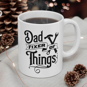 May include: White ceramic coffee mug with black text that reads "Dad Fixer of Things" with a wrench and hammer graphic.