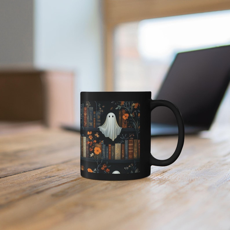 A black mug with a Halloween-themed design featuring a ghost surrounded by books and flowers on a wooden table.