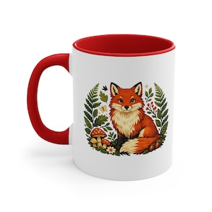 Puede incluir: Una taza de cerámica blanca con asa y borde interior de color rojo brillante. El exterior presenta una colorida ilustración de un zorro sentado entre helechos verdes, pequeños hongos de sombrero rojo, una mariposa amarilla y flores blancas.
