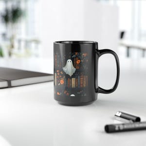 A black mug with a Halloween-themed design featuring a ghost, flowers, and books on a white background.