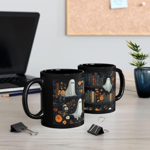 two black mugs with a Halloween-themed design, including ghostly figures and floral elements, placed on a desk with a laptop and office supplies.
