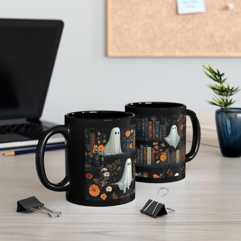 two black mugs with a Halloween-themed design, including ghostly figures and floral elements, placed on a desk with a laptop and office supplies.