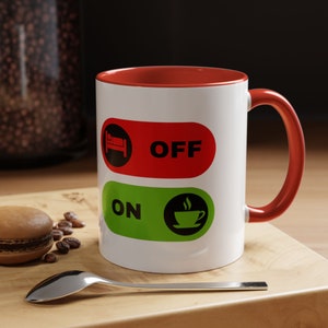 May include: White ceramic coffee mug with a red rim and handle. The mug has a graphic of two toggle switches. The top switch is red and says "OFF" with a sleeping person icon. The bottom switch is green and says "ON" with a coffee cup icon.