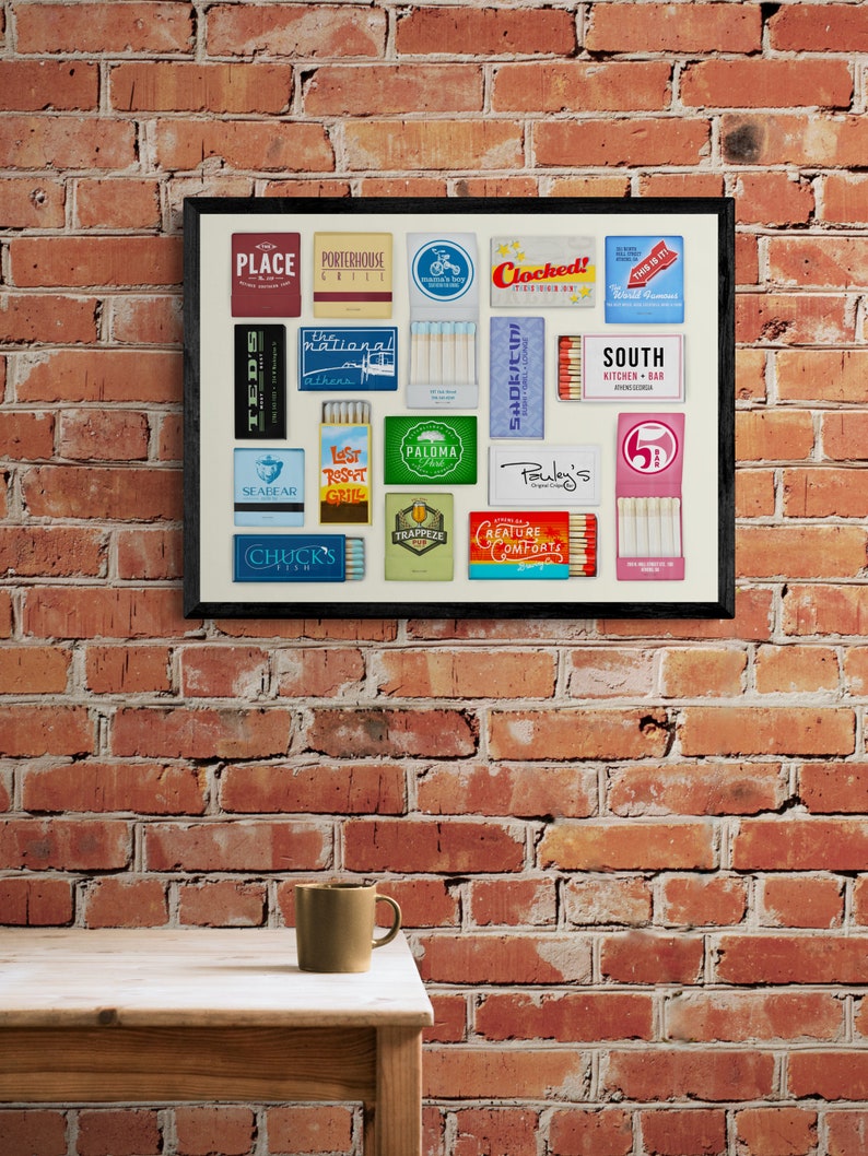 May include: A framed collage of matchbox labels from various restaurants and bars. The labels feature colorful designs and text, including names like "Clocked!", "South Kitchen + Bar", "Pauley's", and "Seabear".