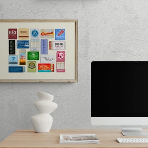 May include: A framed collage of vintage matchbook covers featuring various restaurant and bar names, including "Place", "Cocktails", "South Kitchen & Bar", and "Palma". The collage is displayed on a white wall above a desk with a computer monitor and a white ceramic vase.