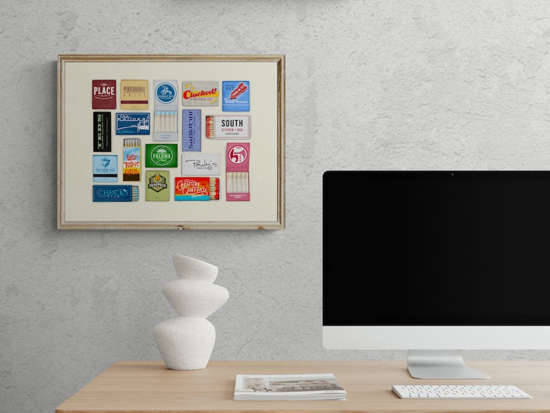 May include: A framed collage of vintage matchbook covers featuring various restaurant and bar names, including "Place", "Cocktails", "South Kitchen & Bar", and "Palma". The collage is displayed on a white wall above a desk with a computer monitor and a white ceramic vase.